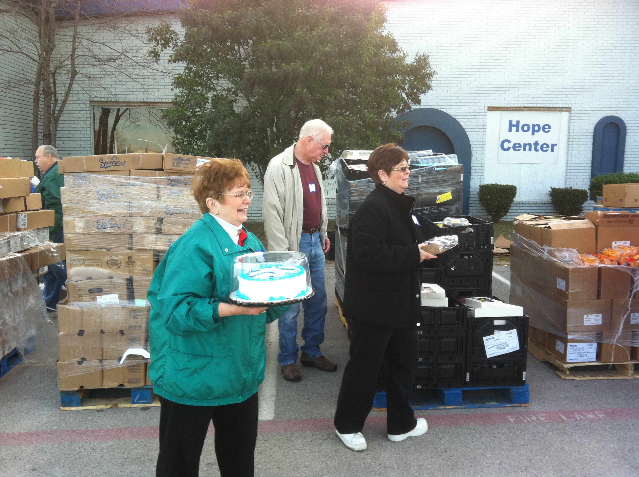 Volunteers outdoors by pallets of food boxes at Hope Center; woman holds a frosted cake