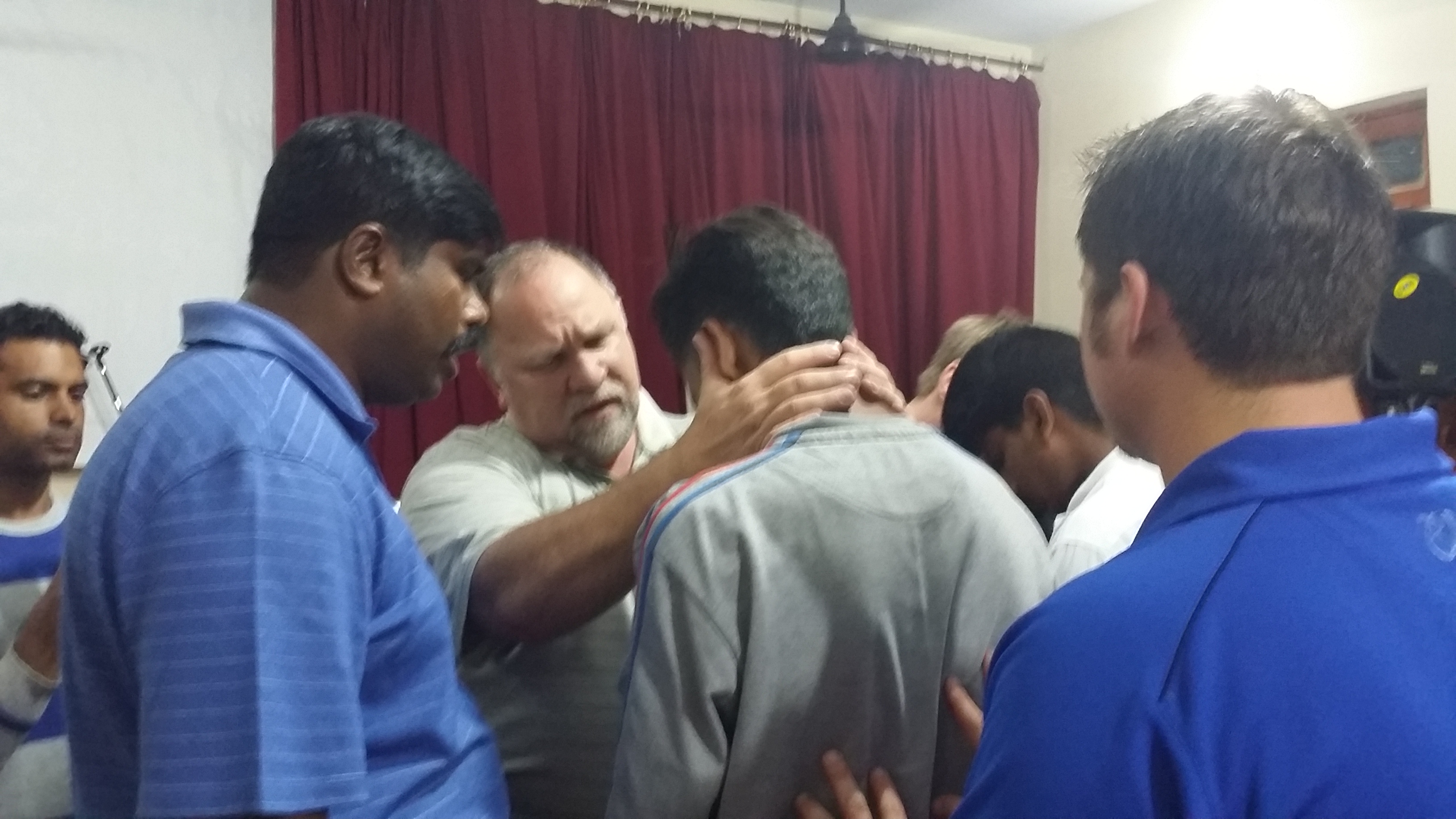 Indoor prayer laying-on-of-hands with South Asian men; red curtain backdrop