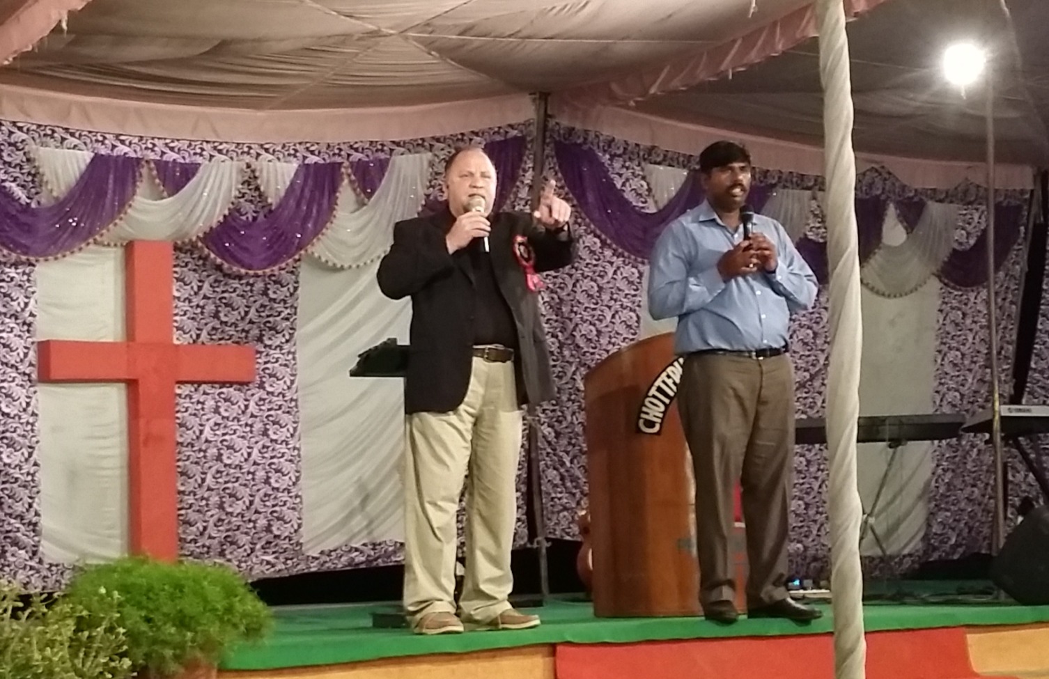 Two men on tent stage with large red cross
