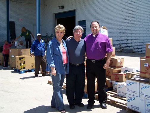 Three adults smiling beside Sysco food boxes; man in purple clerical shirt