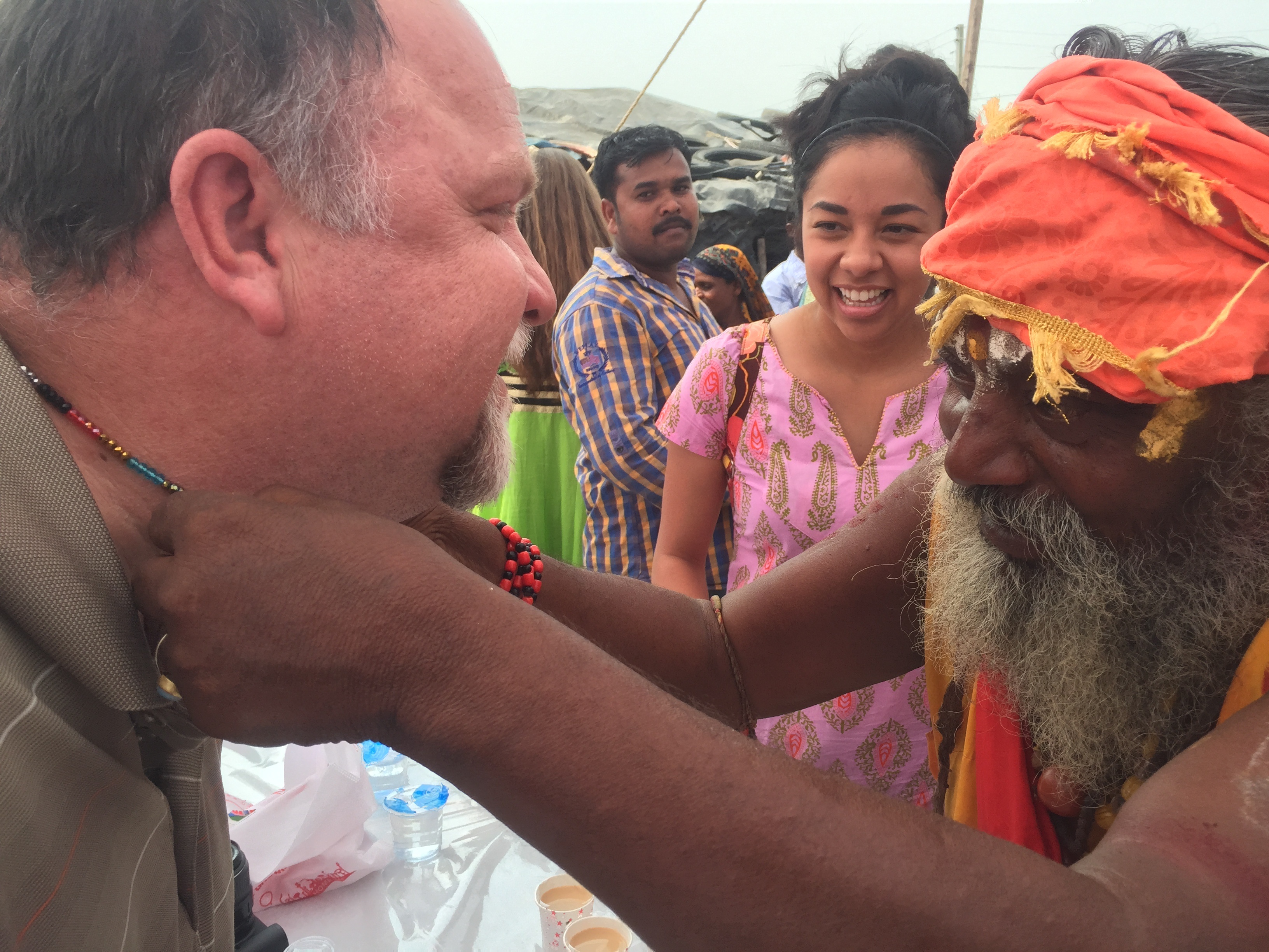 Elderly Indian man in orange turban places beaded necklace on a man; woman in pink kurta smiles